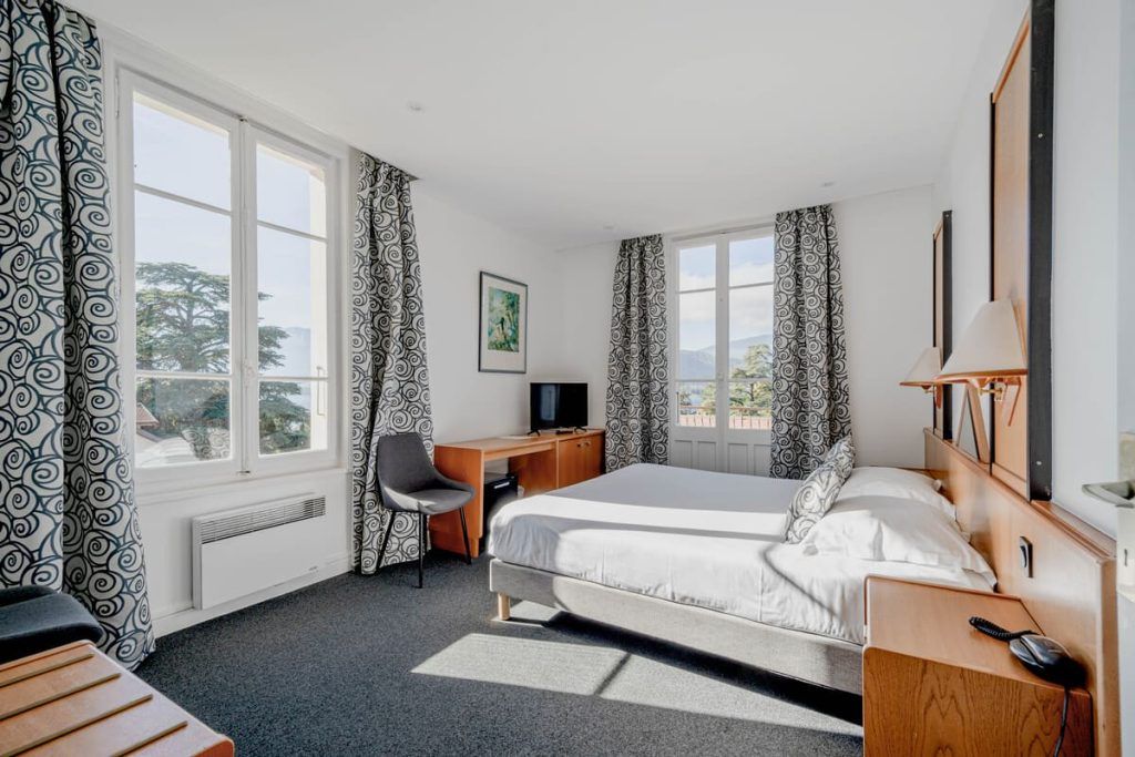 Chambre moderne et lumineuse avec un lit, un bureau, une chaise et de grandes fenêtres avec des rideaux à motifs.