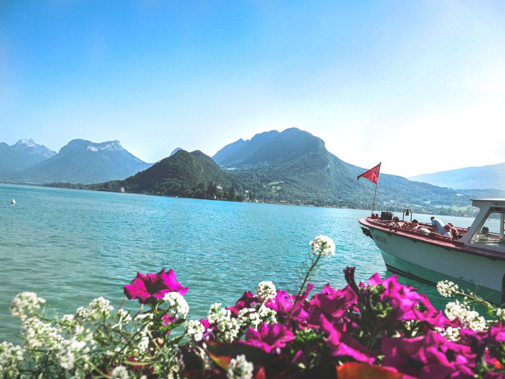 Des fleurs éclatantes surplombent un lac turquoise, avec des montagnes et un bateau sous un ciel dégagé.