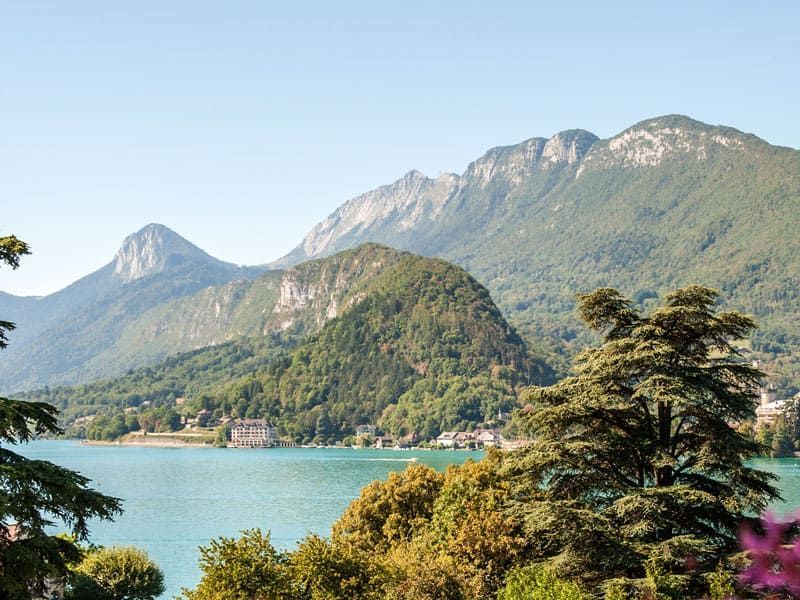 Lac de montagne aux eaux bleues, collines boisées et sommets enneigés sous un ciel dégagé.