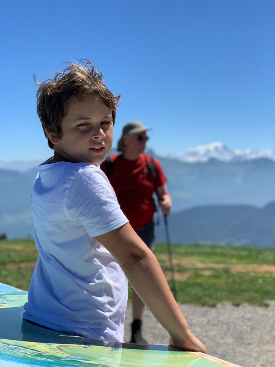 Un enfant en chemise blanche regarde l'objectif depuis un belvédère ensoleillé en montagne, avec une autre personne et des sommets enneigés en arrière-plan.