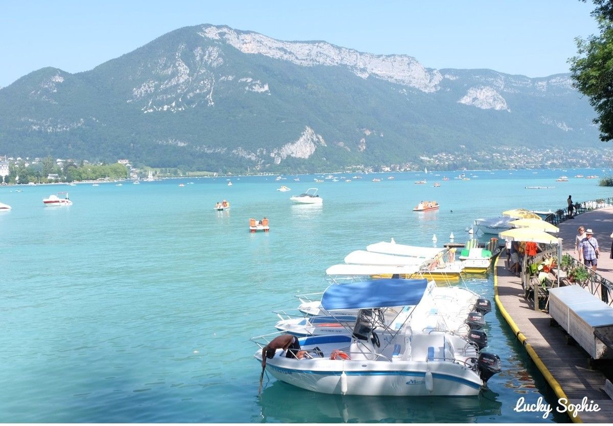 Lac turquoise avec des bateaux, une rive aménagée et des montagnes sous un ciel bleu limpide.