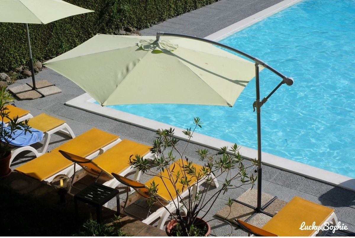 Chaises longues au bord de la piscine, sous un grand parasol vert pâle, près d'une eau d'un bleu éclatant.