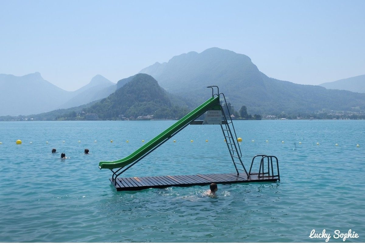 Toboggan aquatique vert flottant sur une plateforme au milieu d'un lac bleu calme, avec des montagnes en arrière-plan.