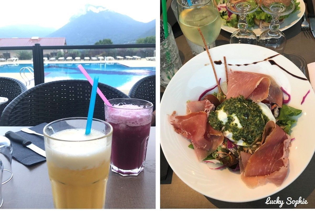 Table de café en terrasse avec boissons colorées et salade, donnant sur une piscine et les montagnes.