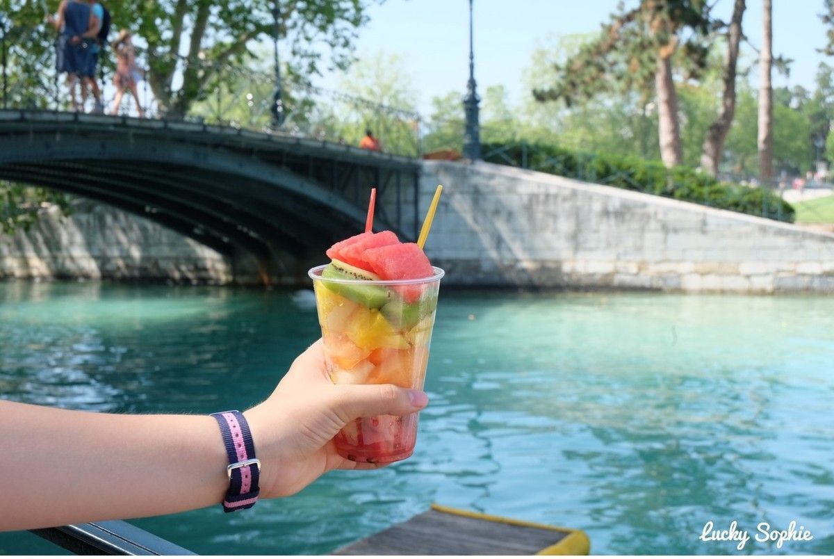 Une main tenant un jus de fruits coloré au bord d'une rivière turquoise, sous un pont de pierre