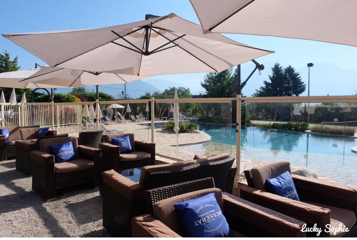 Terrasse aménagée avec chaises en osier, coussins bleus, parasols blancs et piscine au-delà d'une balustrade en verre.