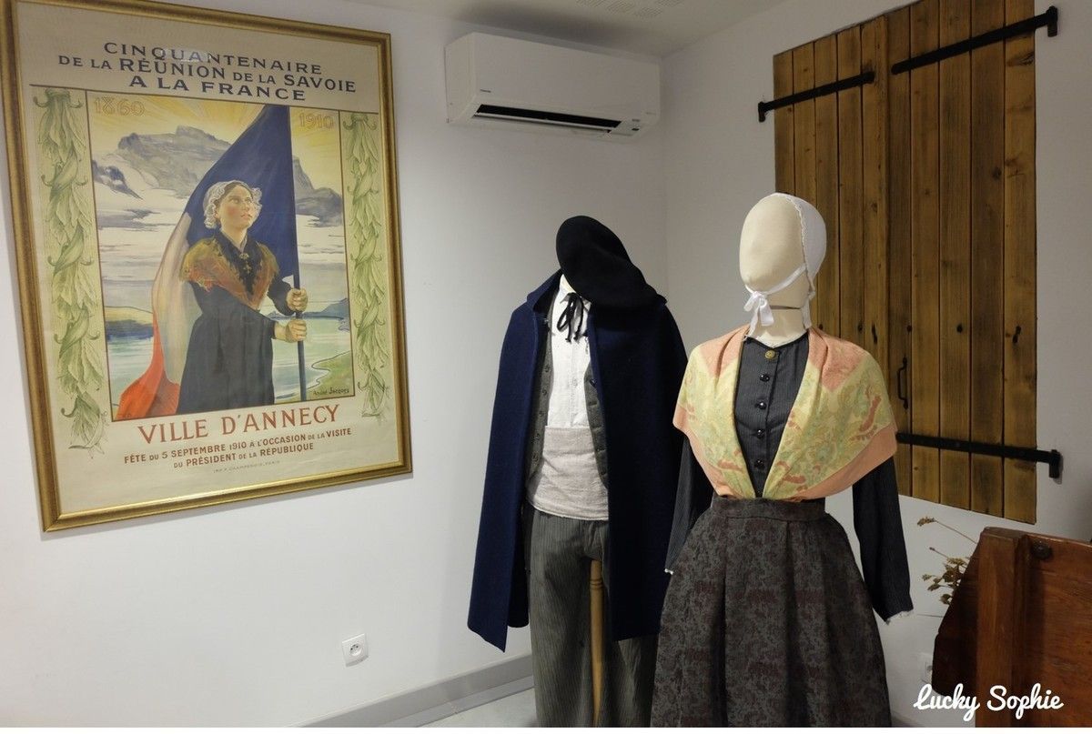 Exposition muséale avec deux mannequins en costumes d'époque à côté d'une grande affiche encadrée et de volets en bois.