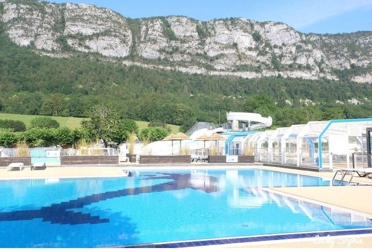 Piscine extérieure aux eaux bleues, adossée à une montagne rocheuse et bordée d'arbres, sous un ciel dégagé.