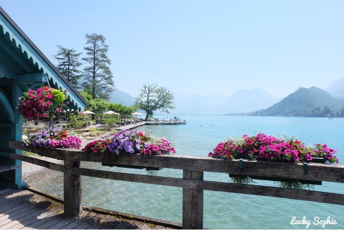 Des jardinières fleuries sur une terrasse au bord d'un lac aux eaux turquoise, avec des montagnes à l'horizon.