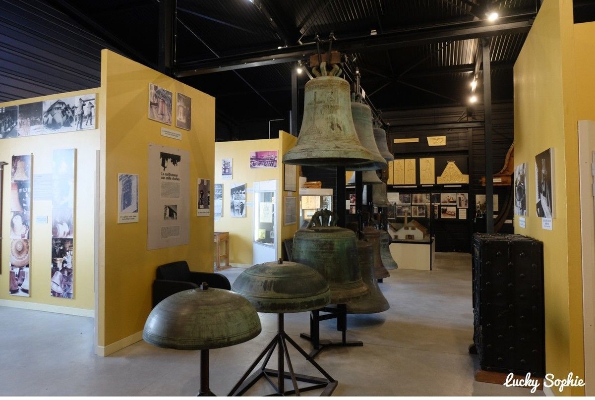 Exposition muséale avec de grandes cloches et des panneaux d'exposition dans une galerie intérieure faiblement éclairée