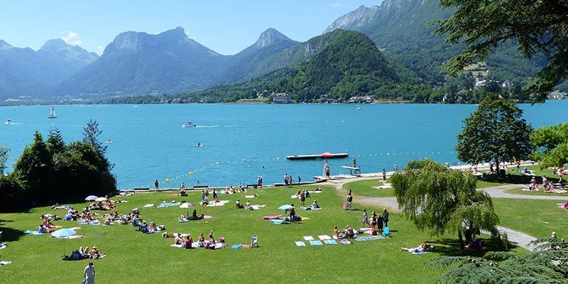 Des personnes se détendent sur une pelouse verdoyante au bord d'un lac d'un bleu éclatant, entourées de montagnes, par une journée ensoleillée.