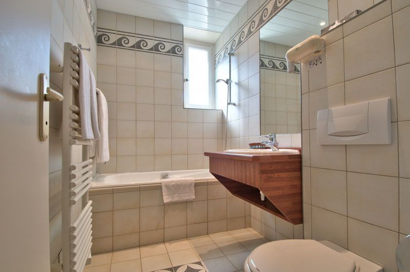 Petite salle de bains carrelée avec baignoire, meuble-lavabo en bois, toilettes et éclairage par fenêtre.