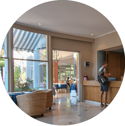 Hall d'entrée lumineux d'un café avec fauteuils en osier, portes vitrées, auvent rayé et une personne au comptoir