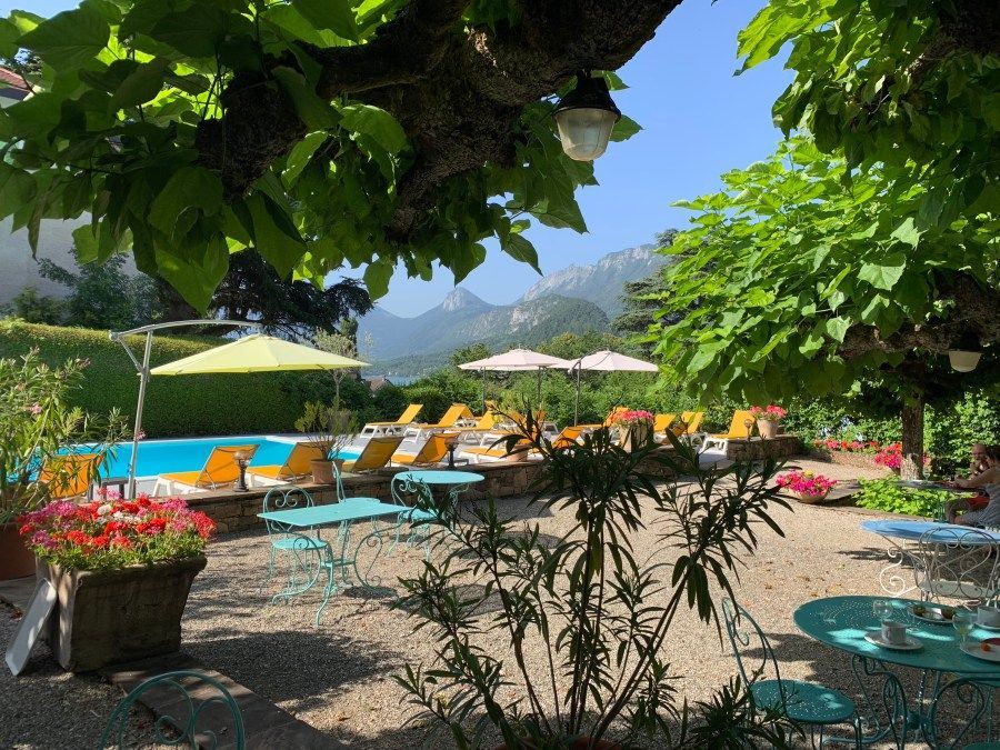 Terrasse ensoleillée au bord de la piscine avec chaises longues turquoise, parasols blancs, fleurs et vue sur les montagnes