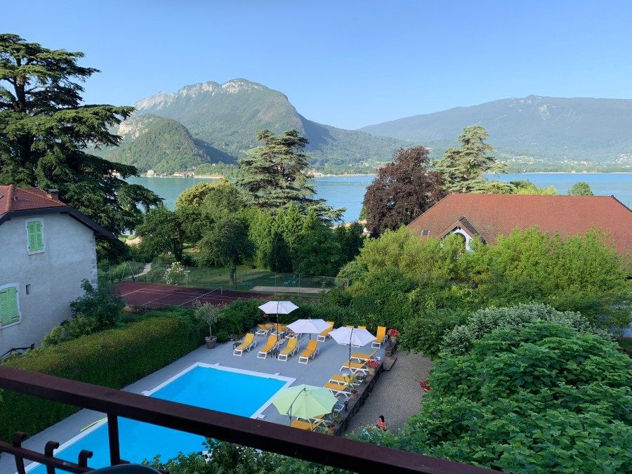 Vue depuis le balcon sur une piscine, des jardins, des bâtiments aux toits rouges et un lac avec des montagnes à l'horizon.