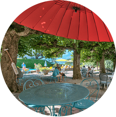 Terrasse ombragée de café avec parasol rouge, arbres verts et tables en métal bleu donnant sur un parc