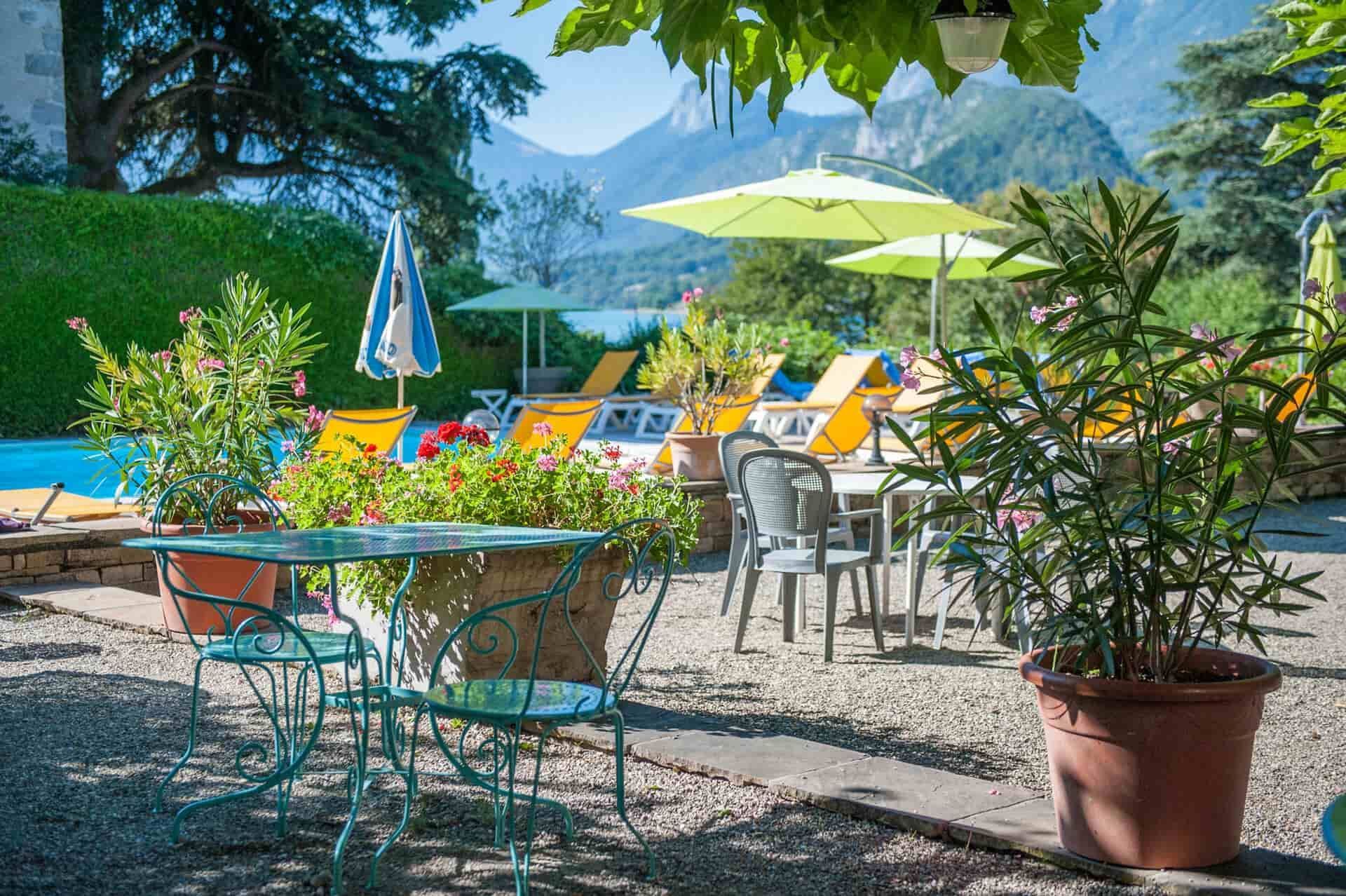 Terrasse ensoleillée d'un café au bord d'un lac, avec chaises colorées, parasols et plantes en pot, et les montagnes en arrière-plan.