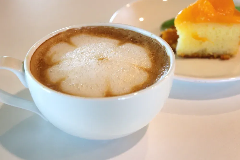 Una taza blanca de café con leche decorada con espuma en forma de flor, junto a una porción de pastel con cítricos