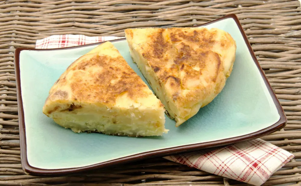 Una taza blanca de café con leche decorada con espuma en forma de flor, junto a una porción de pastel con cítricos