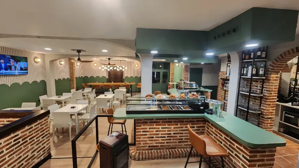Una cafetería rústica con barra de ladrillo, detalles en verde, mesas, sillas y estanterías repletas de botellas.