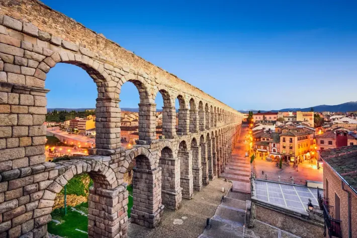 Antiguo acueducto romano en Segovia, España, se arquea sobre una ciudad al anochecer, con edificios y farolas.