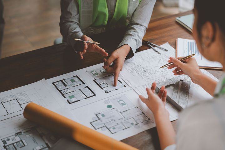 Des personnes assises à une table examinent des plans architecturaux, pointant du doigt et discutant des croquis.