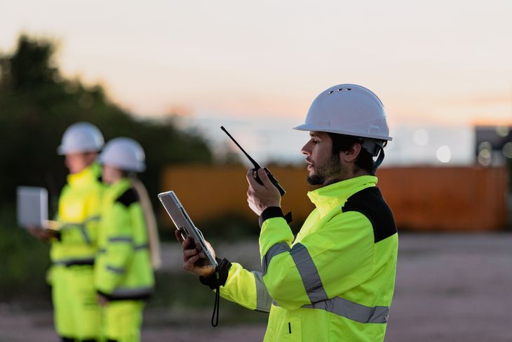 Des ouvriers du bâtiment, vêtus de gilets de sécurité et de casques de chantier, communiquent par talkie-walkie à l'extérieur au crépuscule.