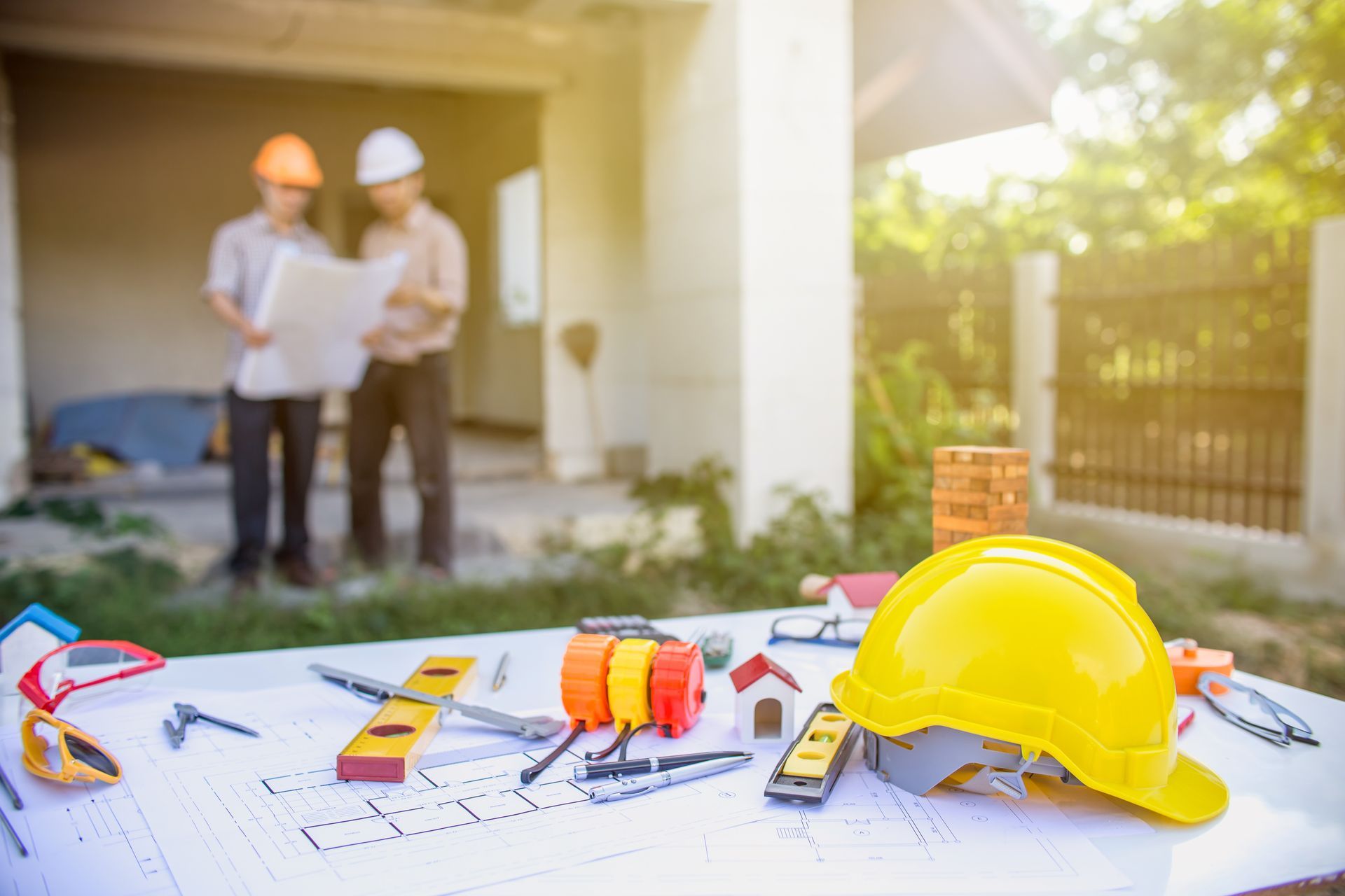 Plans de construction avec un casque et des outils au premier plan ; deux ouvriers examinent des plans sur un chantier de maison.