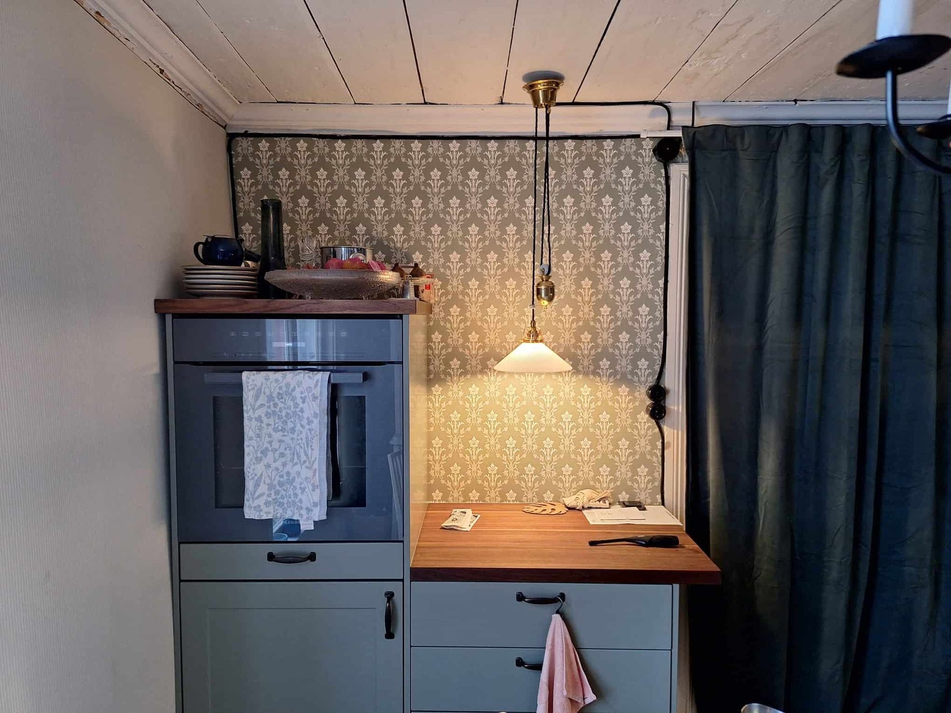 Small kitchen with an oven and cabinets in shades of green, countertop, wallpaper, and a hanging light.