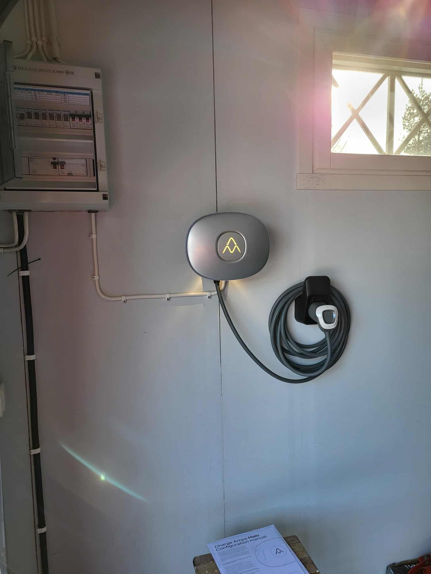 Electric vehicle charger and control panel mounted on a white wall next to a window. A charging cable is coiled below the charger.