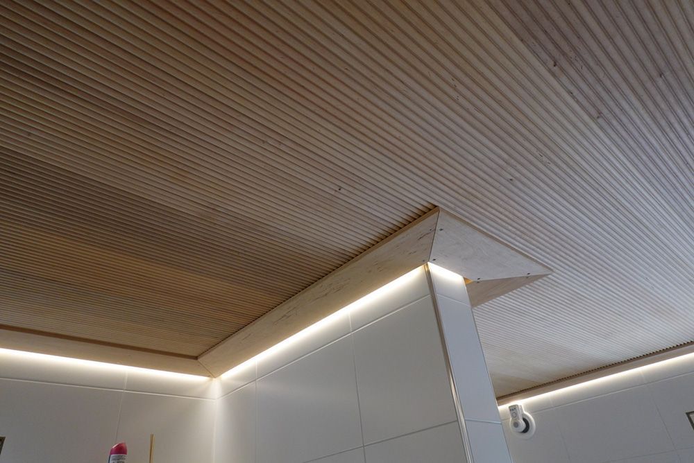 Wooden slatted ceiling with indirect lighting and a white wall/pillar. The lighting is creating a warm ambiance.