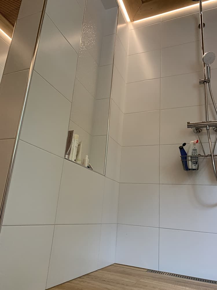 White-tiled shower with a recessed shelf holding toiletries. Chrome shower fixtures contrast against the white walls and wood-look flooring.