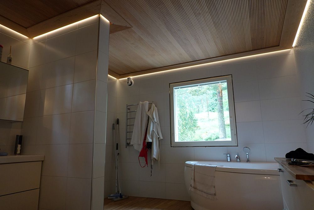 Bathroom with wood ceiling and LED strip lighting. A window overlooks greenery, with a bathtub and white tiled walls.
