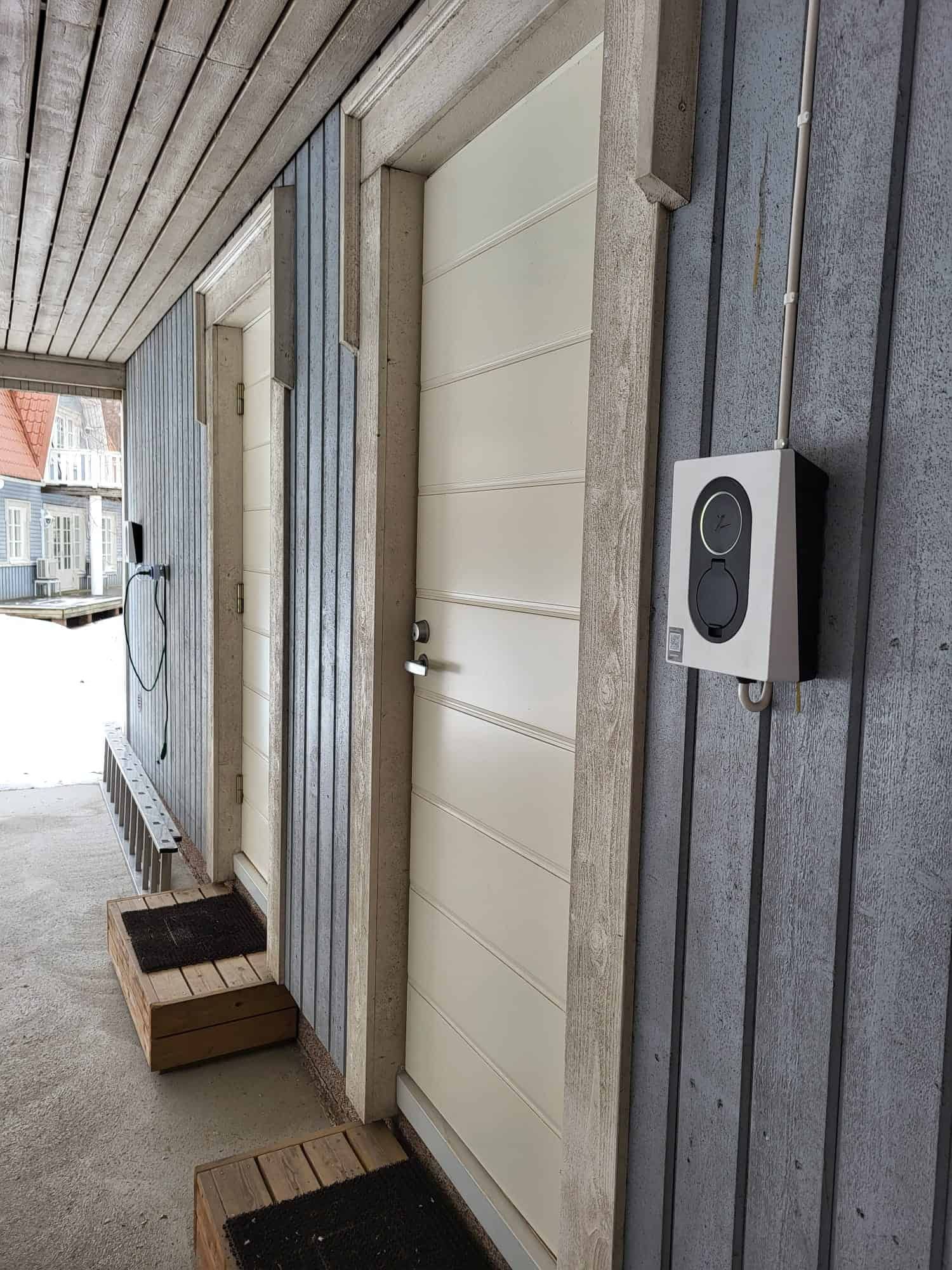 Exterior doors and a charging station on a gray building with wood trim. Two doors are visible, with welcome mats on wooden steps.