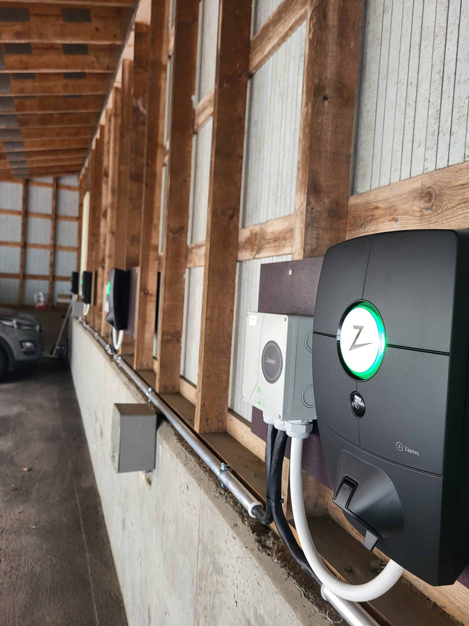 Several black EV chargers mounted on a concrete wall inside a wooden-framed structure. Green lights indicate charging status.