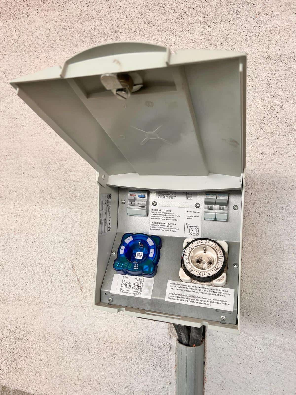 Outdoor electrical outlet box on a wall, open to reveal a blue and a white outlet. A gray lid is propped open above them.