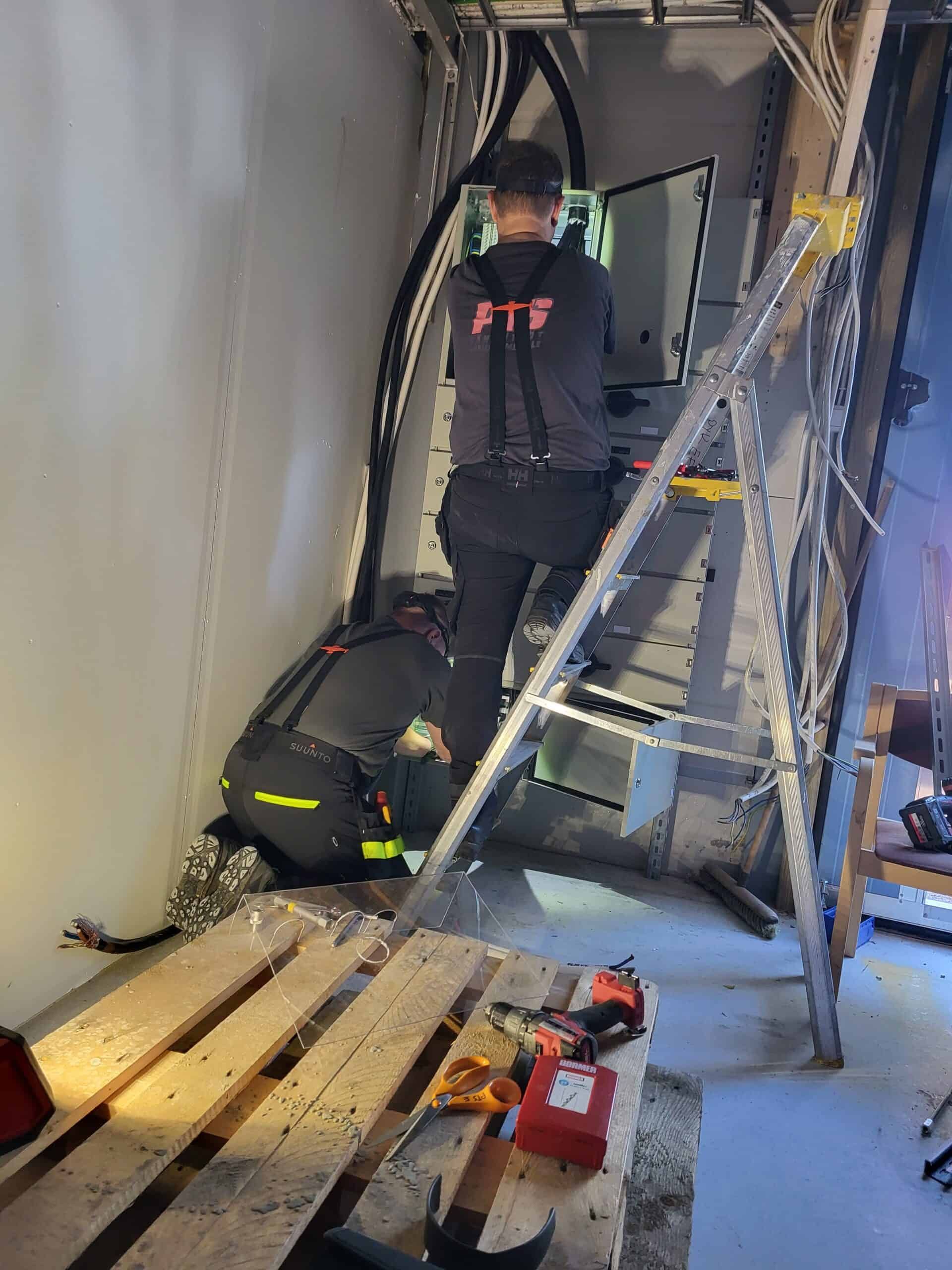A person in a black shirt and pants, on a ladder, working on electrical wiring in a metal-walled space. Tools and pallets are on the floor.