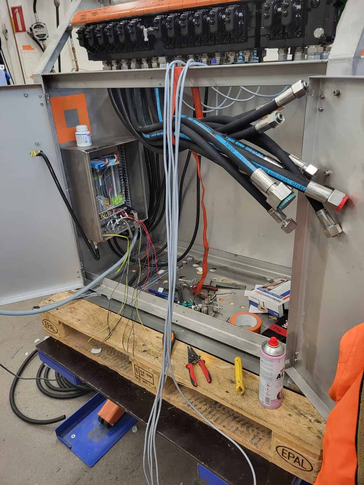 Wiring work in progress, with an open metal cabinet, tools, and hydraulic lines. Wires dangle from the upper portion of the cabinet.