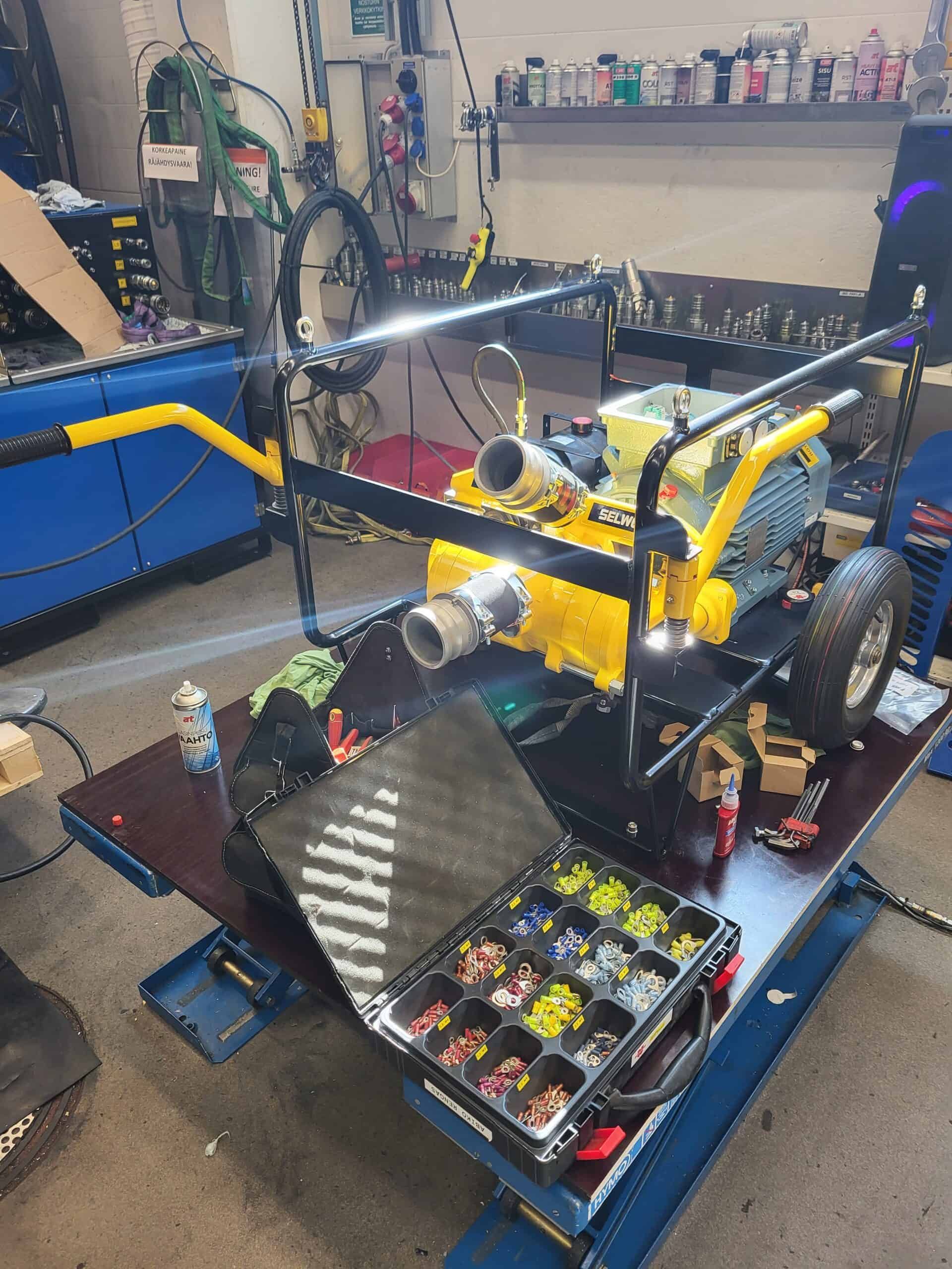 A yellow and black industrial machine on a lift table, with a case of colored parts in front. Workshop setting.