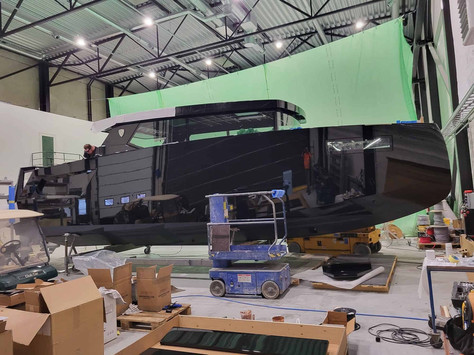 A large black yacht being worked on inside a warehouse with a green screen background. Tools and materials surround it.