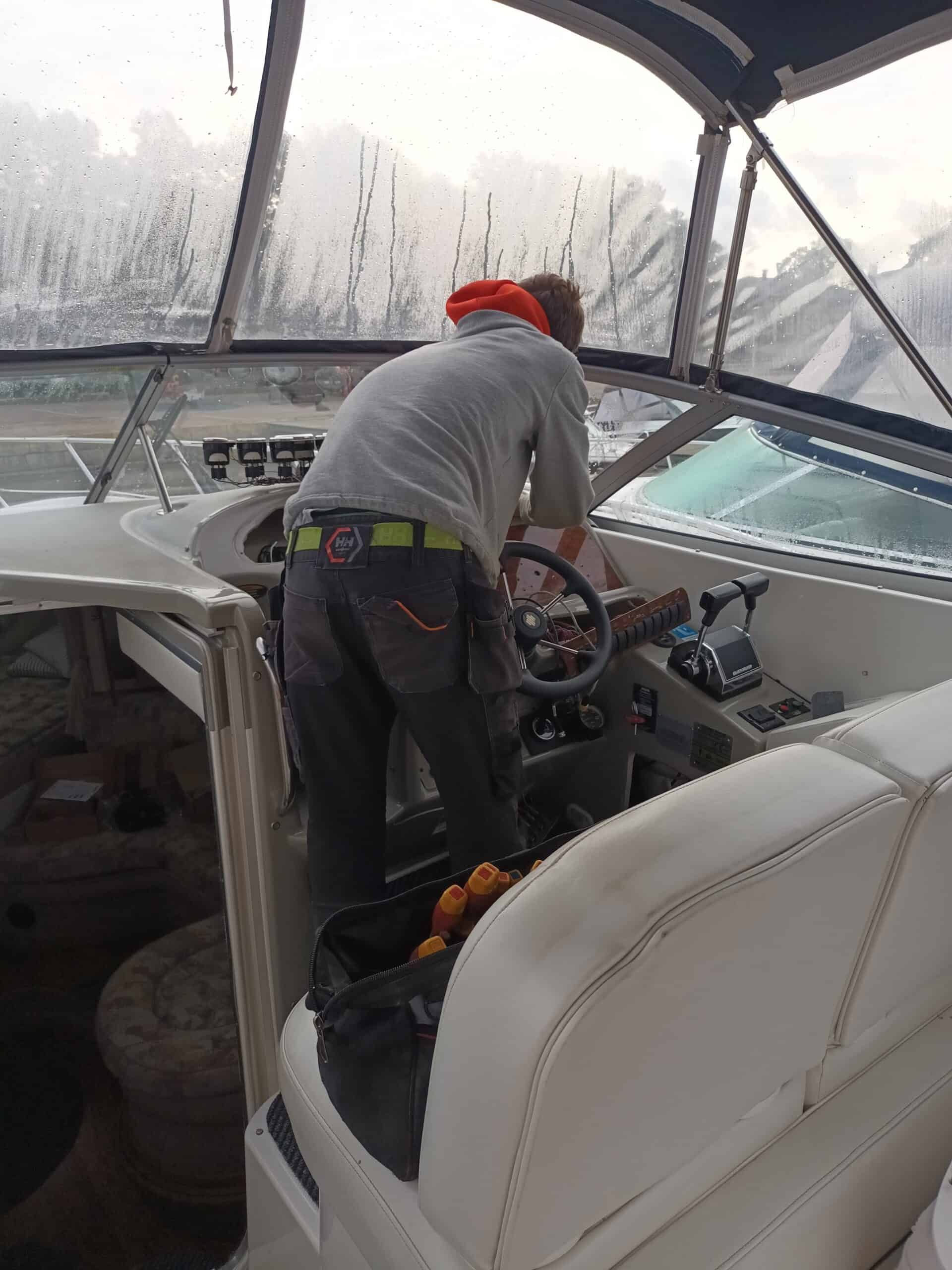 A person in gray clothing and an orange hat operates a boat's control panel, inside the boat's cabin.