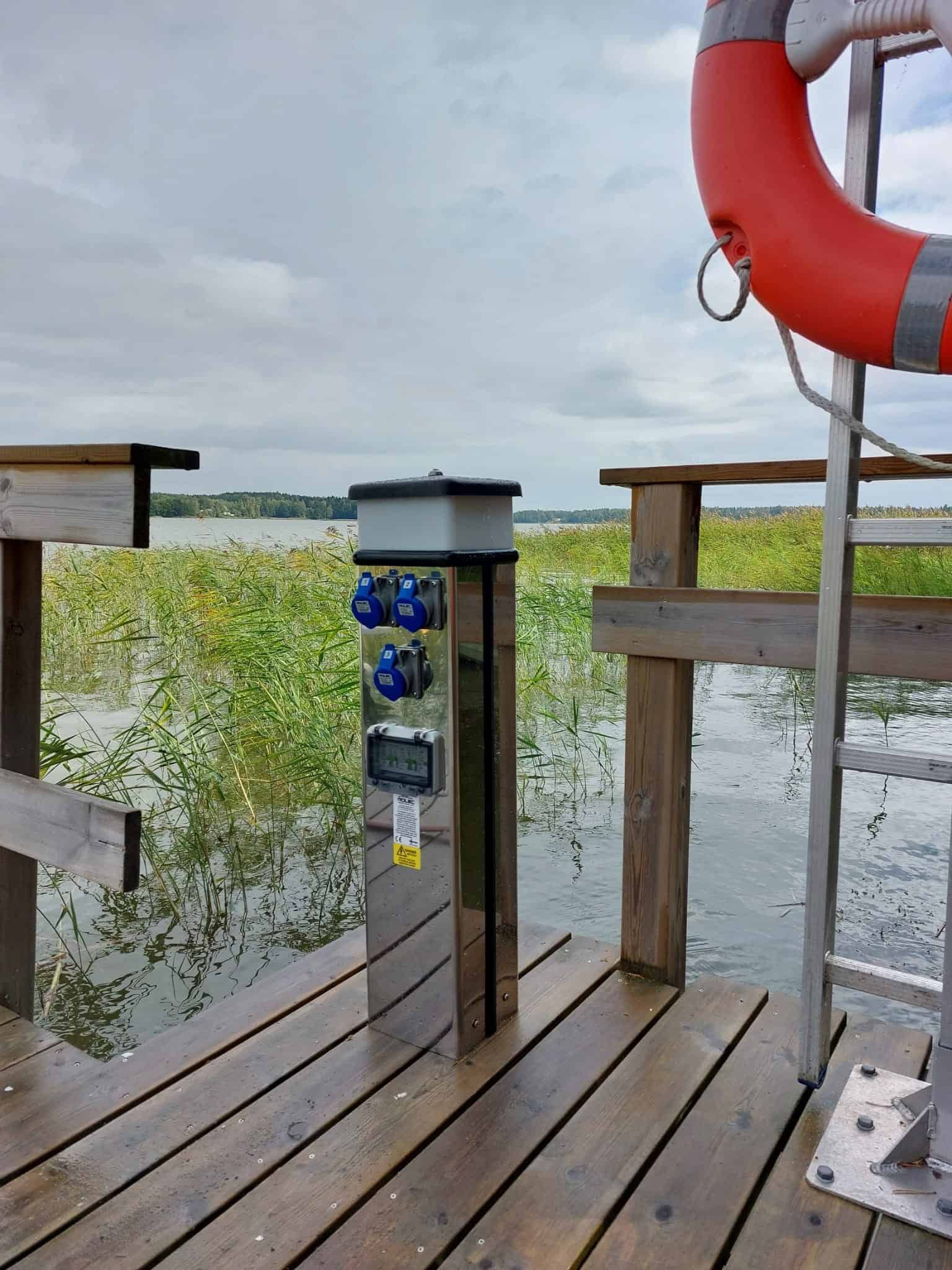 Wooden dock with electrical outlets and a life preserver overlooking a body of water and reeds under a cloudy sky.