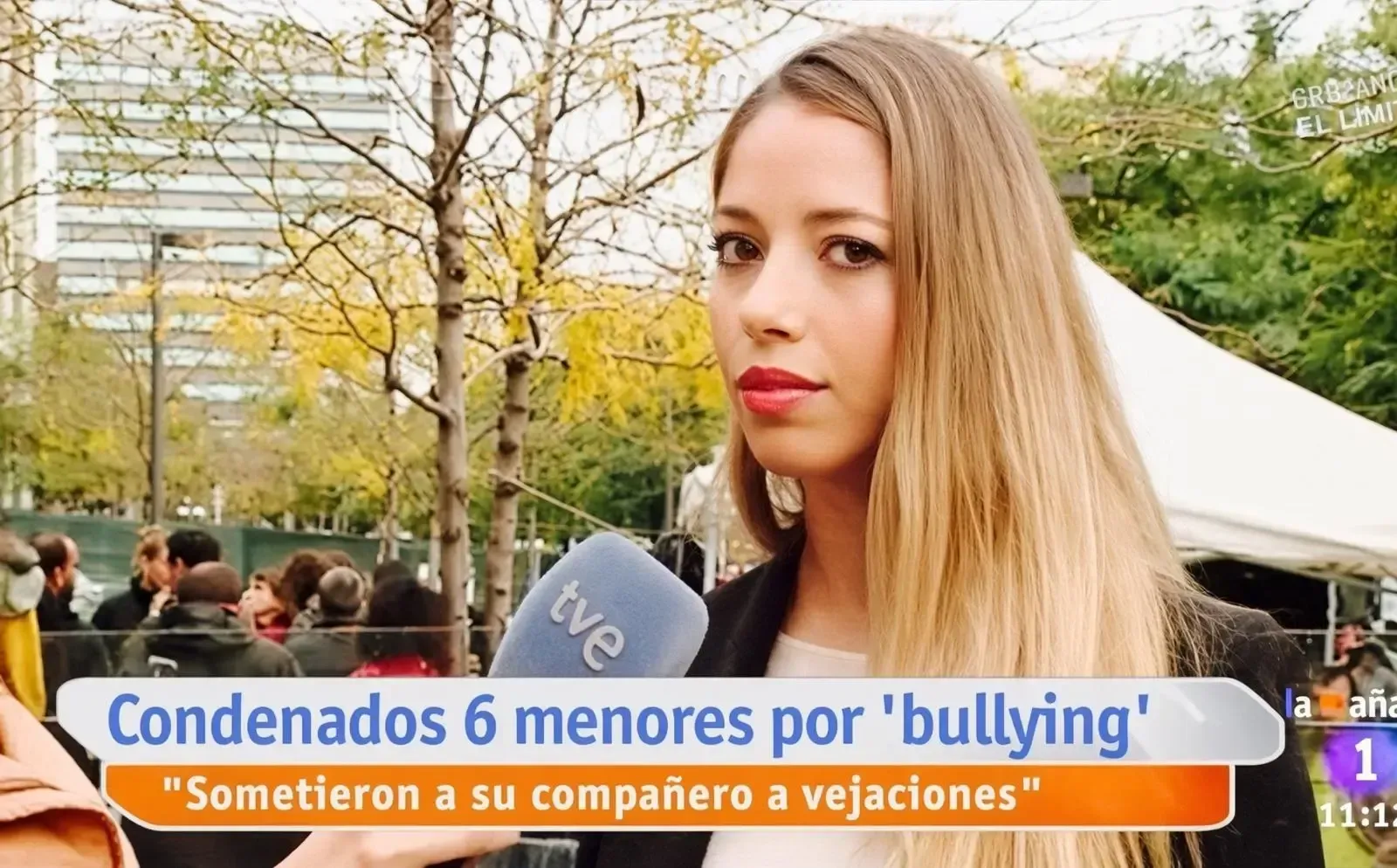 Una mujer de pelo largo y rubio está siendo entrevistada al aire libre, con un cartel de noticias en español sobre una sentencia por acoso escolar.