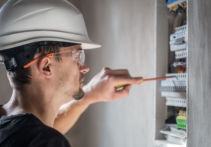 Electricista con casco blanco y gafas de seguridad trabajando en un panel de disyuntores.