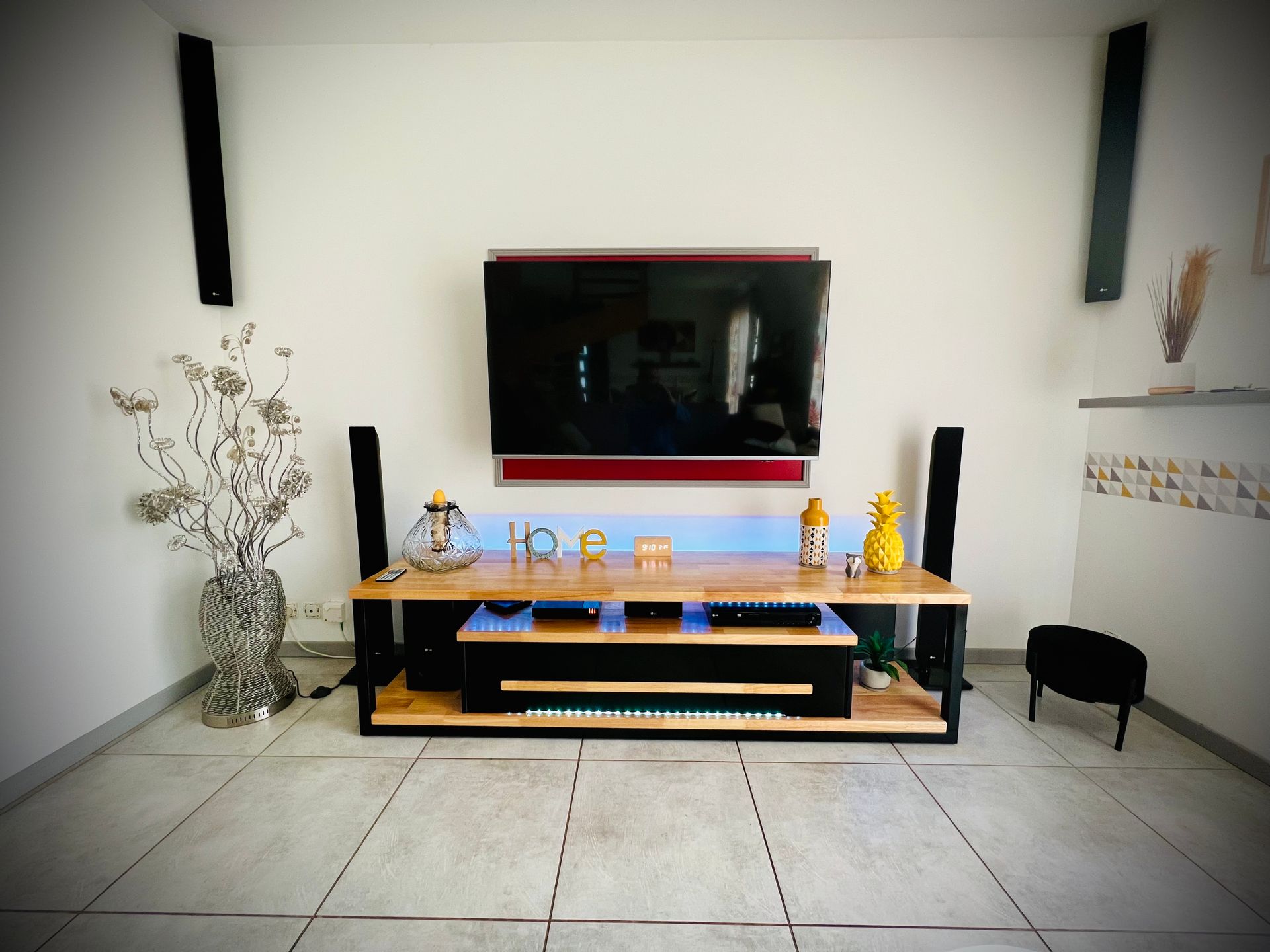 Salon minimaliste avec télévision murale, console basse en bois, vase de branches et tabouret noir.