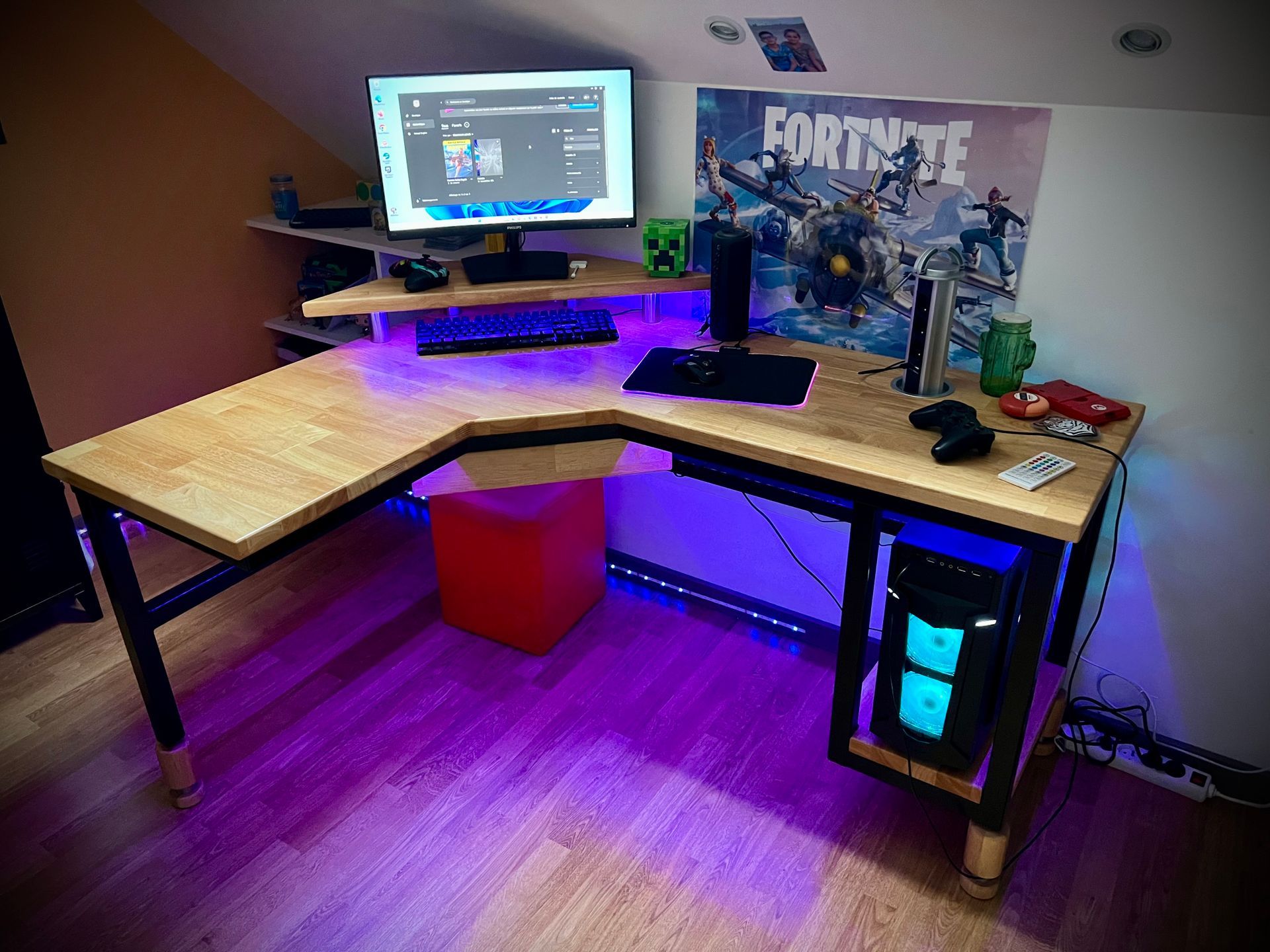 Bureau gaming en forme de L avec écran, éclairage LED violet, poster Fortnite et tour PC en dessous.