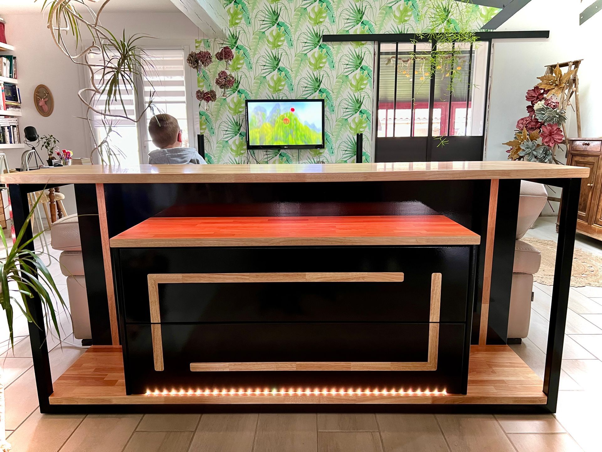 Banc d'intérieur moderne avec structure noire, coussin orange et bande LED, dans une pièce lumineuse remplie de plantes.