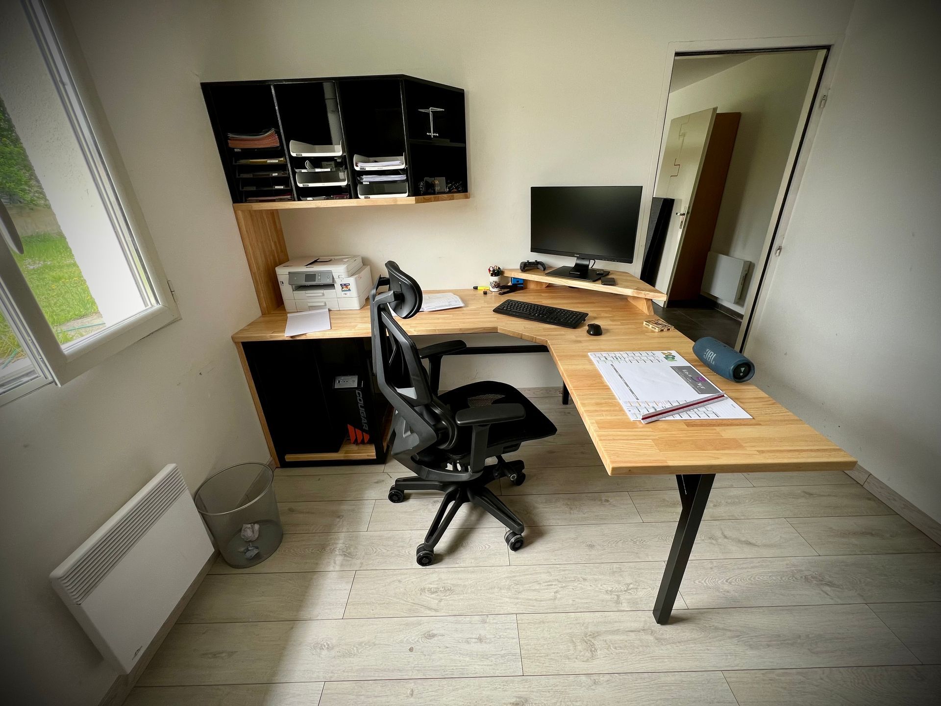 Petit bureau avec bureau, chaise, armoires murales, imprimante et grand écran près d'une fenêtre.
