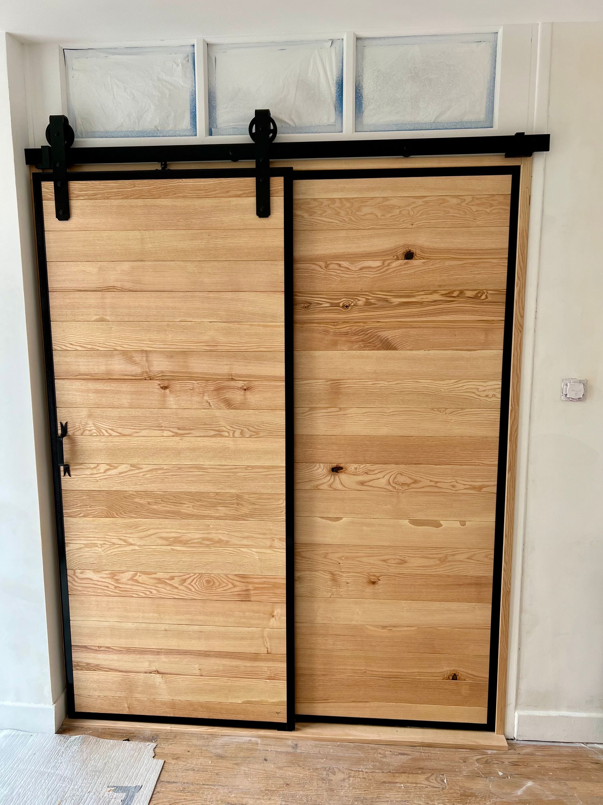 Portes coulissantes en bois avec quincaillerie noire sous trois fenêtres dépolies sur un mur blanc.