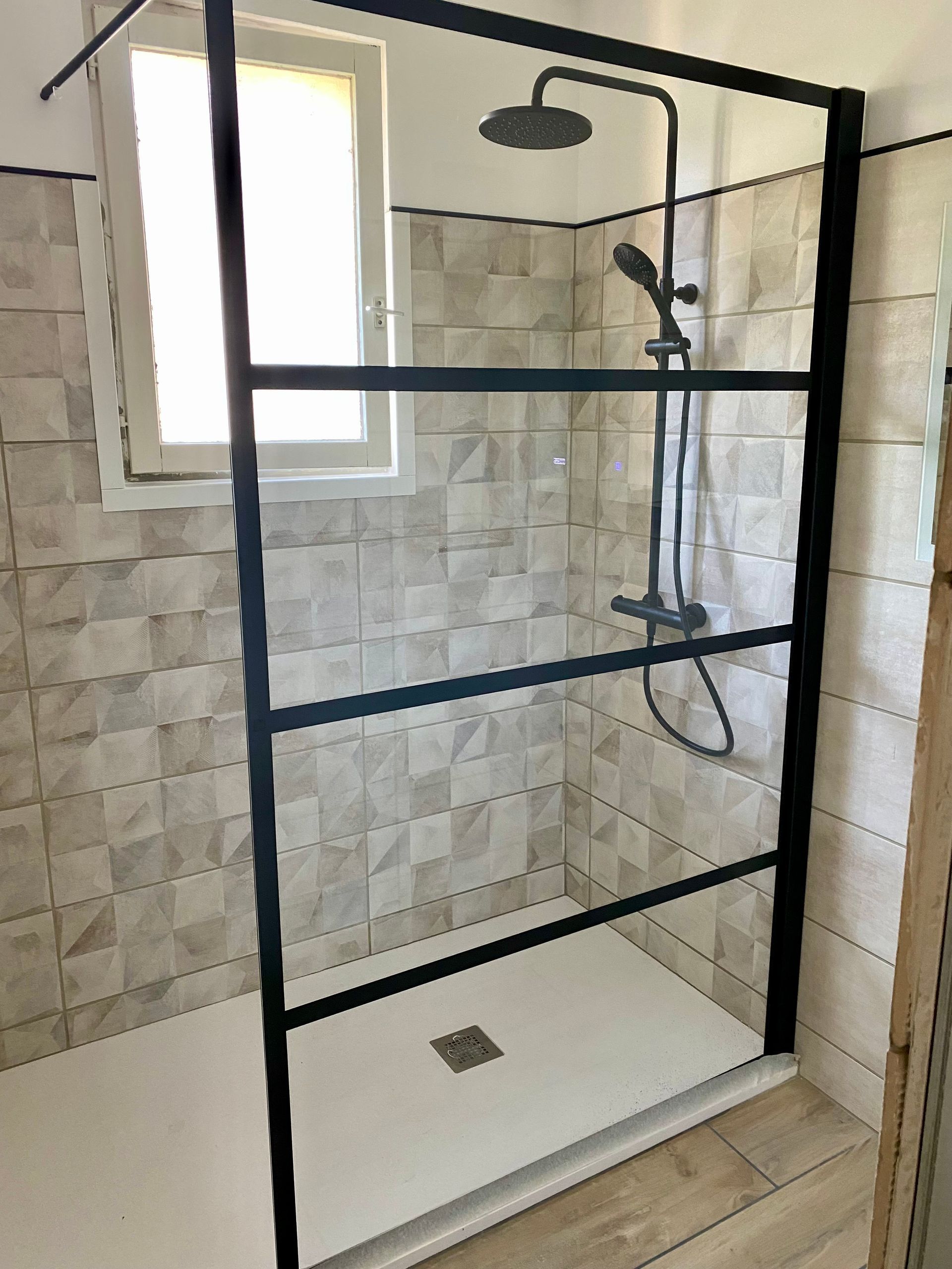 Douche de salle de bains avec paroi de douche en verre à cadre noir, murs carrelés et pommeau de douche sur socle blanc.
